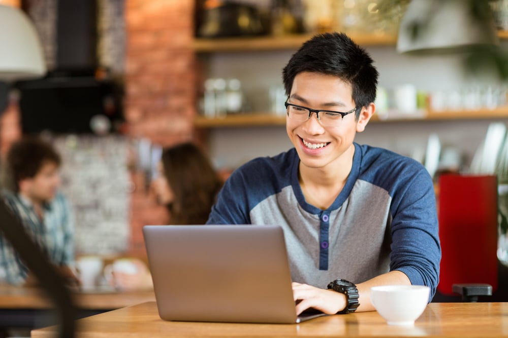 Happy cheerful young asian male in glasses smiling and using laptop in cafe Happy cheerful young asian male in glasses smiling and using laptop in cafe
