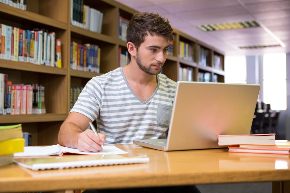 Male student sitting at a desk in the library working on his laptop.
